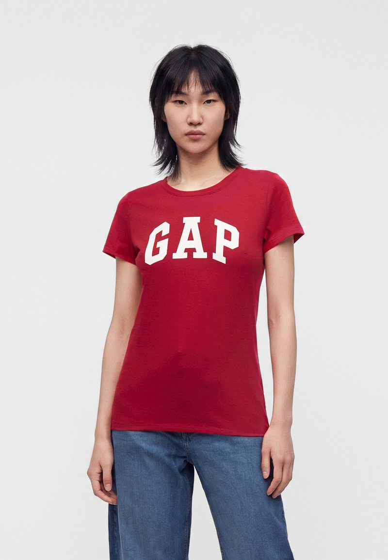 Personne debout, portant un T-shirt rouge à manches courtes avec de grandes lettres blanches GAP et un jean bleu sur un fond uni clair.