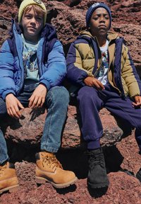 Due ragazzi sono seduti sulle rocce. Uno indossa un giubbotto a vento blu, un maglione azzurro chiaro e jeans con stivali gialli. L'altro indossa un giubbotto a vento verde e blu, pantaloni scuri e stivali neri.
