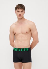 Svarte boksershorts med en bred, elastisk grønn midje som har "CALVIN KLEIN" i fet skrift; glatt stoff og tettsittende design.