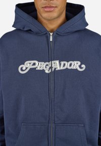 Marineblå zip-up hoodie lavet af bomuldsblanding, med en frontlomme og hvid "PEGADOR" tekstlogo på tværs af brystet. Blød tekstur.