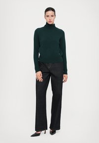Donkergroen gebreid coltrui gedragen met hoge taille donkerdenim jeans en spitse zwarte hakken, wat een gestructureerde silhouet creëert.