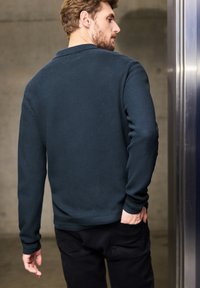 Navyfarbener Pullover mit Struktur und Kragen, mit langen Ärmeln und gerippten Bündchen, kombiniert mit schwarzen Hosen. Der Hintergrund ist eine Betonwand.