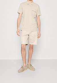 Polo beige avec une poche, associé à un short beige et des chaussures en suède gris clair. La tenue présente une coupe décontractée et un design simple.