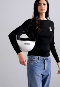 Hvid hjelm med sorte accenter og ordet "MOSCHINO" trykt på, holdt under armen af en person i en sort ribbet sweater og blå jeans.