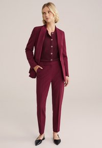 Costume bordeaux ajusté comprenant une veste cintrée et un pantalon fuselé, avec un haut texturé bordeaux et des boutons ronds. Chaussures plates noires pointues.
