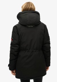 Zwarte, gewatteerde parka jas met een grote capuchon, tailleriem en Superdry-logo op de achterkant en mouw. Heeft een gladde, matte textuur.