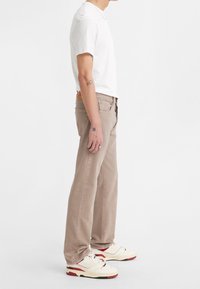 Jeans rectos de color beige claro, hechos de mezclilla. Usados con una camiseta blanca ajustada y zapatillas beige con acentos rojos. Vista de perfil lateral.