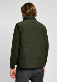 Gilet imbottito verde scuro con colletto a coste, caratterizzato da un motivo trapuntato sulla parte superiore della schiena e una sezione inferiore liscia.