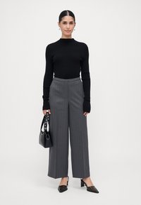 Vrouw gekleed in een zwarte lange mouwen top, grijze streepjaspantalon met brede pijpen, zwarte hakken, en een zwarte handtas, staande tegen een effen achtergrond.