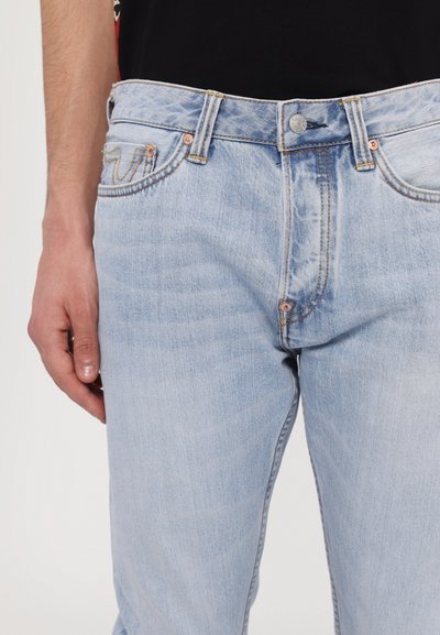 Jean en denim bleu clair avec une coupe slim, présentant une fermeture à bouton, cinq poches et des détails de couture subtils. Texture lisse.
