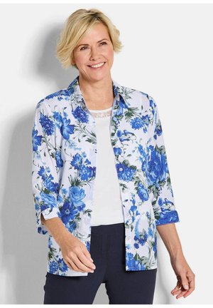 Vrouw die glimlacht, draagt een wit overhemd met een blauw bloemenpatroon over een wit shirt en donkere broek, staand tegen een effen achtergrond.