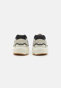 Sneaker mit einem Obermaterial aus Mesh in Beige und Schwarz, ausgestattet mit einer gepolsterten Sohle, runder Form und einem Branding-Detail an der Rückseite. Die Sohle hat ein Profil aus Gummirubber.