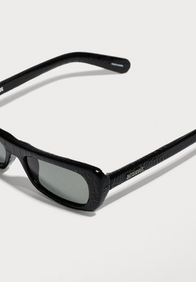 Lunettes de soleil rectangulaires noires avec monture texturée et verres foncés, avec un petit logo "Jacquemus" sur la branche.