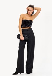 Top crop sin mangas de lentejuelas negras combinado con pantalones negros de talle alto. Accesorios incluyen un bolso de mano de flecos negro y tacones de punta.