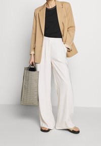 Beige Blazer über einem schwarzen Hemd, weißen weit geschnittenen Hosen und schwarzen Sandalen. Enthält eine karierte Tragetasche mit dunklen Griffen.