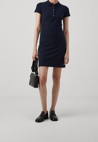 Nainen, jolla on yllään tummansininen polo-mekko, musta käsilaukku kädessä ja mustat loafer-kengät, taustanaan vaalea yksinkertainen tausta.