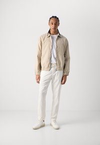 Beige bomberjakke med elastiske manchetter, båret over en hvid skjorte. Kombination med krémfarvede bukser og hvide sneakers. Enkel design, ingen mønstre.