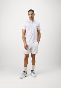 Weißes Polohemd mit blauen Akzenten, kombiniert mit weißen Shorts. Getragen mit weißen Socken und blauen, schwarzen und grünen Sportschuhen.