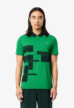 Grøn polo skjorte med sorte geometriske mønstre, korte ærmer og en mørk krave, med et lille logo på venstre bryst.