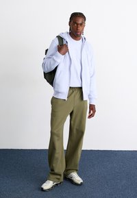 Um hoodie azul claro com fecho éclair sobre uma t-shirt branca, calças de moletão verde-oliva, sapatilhas cinza e pretas, de pé com uma mochila verde.