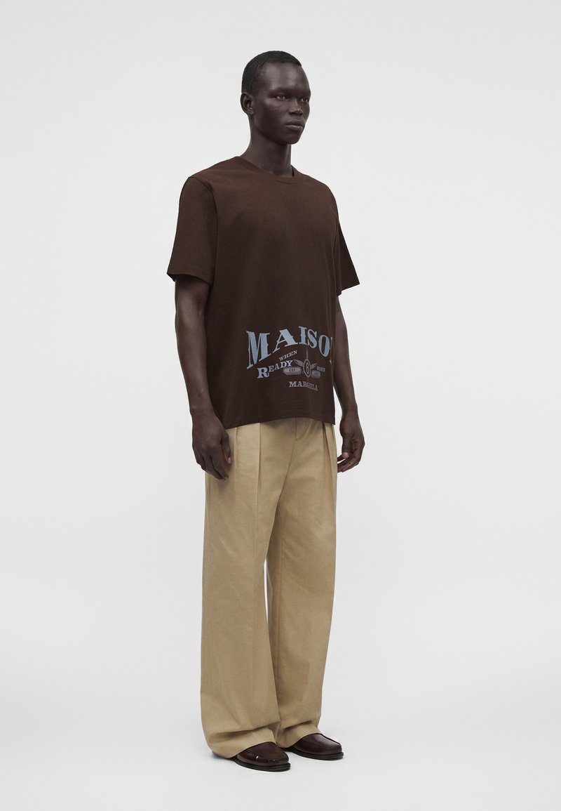 Mann trägt braunes Grafik-T-Shirt, beige Hosen mit weitem Bein und dunkelbraune Schuhe, steht vor einem schlichten hellgrauen Hintergrund.