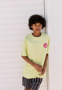 Lichtgele t-shirt met korte mouwen en een ronde halslijn, versierd met een roze grafisch ontwerp op de borst, gecombineerd met zwart-witte gestreepte shorts.