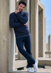 Mann mit Bart, der an einem Betonsäulenrahmen lehnt, trägt einen navyblauen Pullover, dunkle Hosen und weiße Sneakers, draußen unter klarem Himmel.