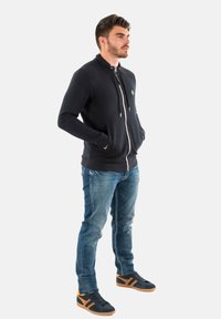Marineblauwe zip-up hoodie met voorzakken, featuring zilverkleurige hardware en een logo, gecombineerd met lichtblauwe jeans en zwart-oranje sneakers.