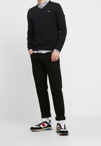 Svart V-ringad tröja, rutigt skjorta under, svarta jeans och svart/vita sneakers med röda accenter. Avslappnad outfit med uppvikt byxben.