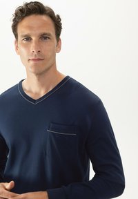Chemise à manches longues bleu marine avec un col en V, une poche poitrine ornée, et une texture de tissu douce. Design épuré et minimaliste.