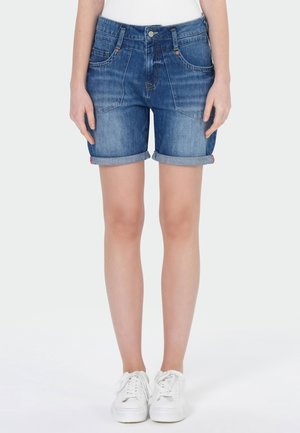 Personne portant un short en jean bleu retroussé et des baskets blanches, debout devant un fond clair uni.
