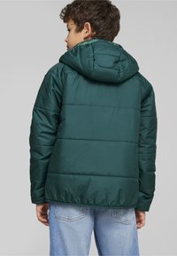 Chaqueta inflable verde con capucha, con un diseño acolchado, textura suave y puños elásticos, combinada con unos vaqueros de color azul claro.