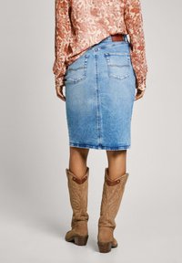 Falda lápiz de mezclilla en azul claro con bolsillos traseros y un dobladillo por debajo de la rodilla, combinada con botas de vaquero de ante color marrón claro con costuras decorativas.