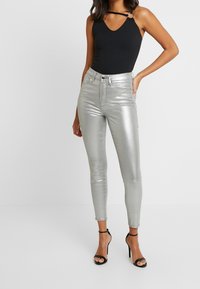 Bodysuit negro con escote en V y detalle de tirantes, combinado con jeans ajustados metálicos plateados, de largo a los tobillos, y sandalias de tacón negras.
