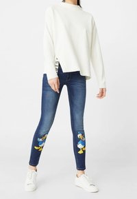 White, long-sleeve top with a side slit. Dark blue jeans featuring Donald Duck prints on the lower legs. Paired with white sneakers.