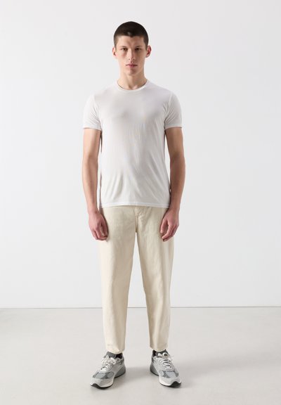 Jeune homme debout, portant un t-shirt blanc ajusté, un pantalon beige, des baskets grises, sur un fond uni clair.