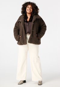 Bruine pluche jas met drukknopen, gedragen over een donker shirt en crèmekleurige wijdelpijpen. De outfit heeft een textuurstof en haklaarzen.