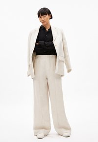 Hellbeige Leinenanzug mit weit geschnittenen Hosen und einem maßgeschneiderten Blazer. Darunter ein schwarzes Hemd mit Knöpfen, getragen mit weißen Sneakers.