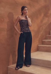 strapless, bedrukt top met geometrische ontwerpen in donkere tinten; losse zwarte broek met zakken; platte zwarte sandalen; natuurlijke, textuur achtergrond.