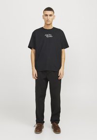 Sort kortærmet t-shirt med "SQUID GAME" grafik i hvid, parret med sorte bukser og brune sko. Enkel, afslappet pasform.
