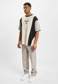 Oversized beige, black, and gray color-blocked t-shirt with short sleeves, featuring a black logo at the chest, paired with cream trousers.