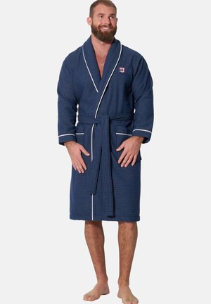 Bearded man standing barefoot wearing a blue striped bathrobe with white trim tied at the waist, smiling and looking to the side.