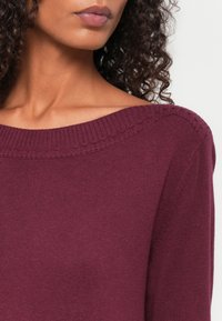 Pull en tricot bordeaux avec un large col côtelé et une texture lisse. Présente des détails d'épaule tombante, montré de la clavicule vers le haut.