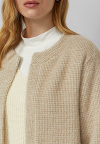 Beige strukturierte Strickjacke mit weichem, flauschigem Finish und dezentem Karo-Muster, getragen über einem cremefarbenen, gerippten Top. Abgerundeter Ausschnitt, offene Vorderseite.