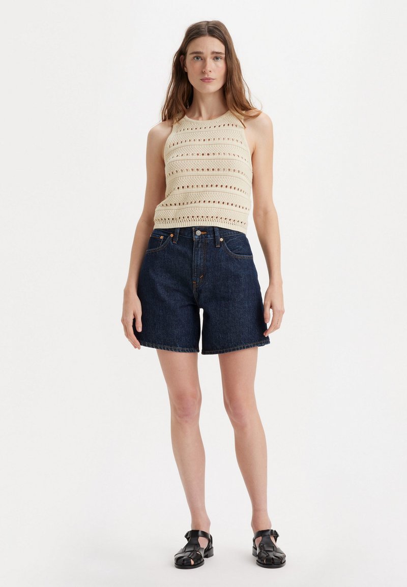 Débardeur en crochet beige avec des motifs horizontaux, associé à un short en denim bleu foncé. Modèle portant des sandales noires, se tenant devant un fond clair.