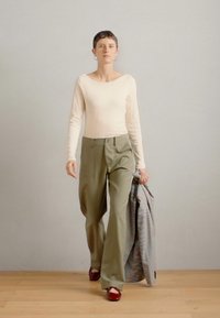 Grietos ilgomis rankovėmis marškiniai, alyvuogių spalvos plačios kojinės kelnės ir raudoni aukštakulniai. Pilkas languotas švarkas užmestas ant vienos rankos. Mediniai grindys, neutralus fonas.