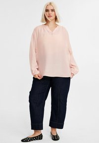 Vaaleanpunainen pusero, jossa on V-pääntie ja pitkät hihat, yhdistetty tummansinisiin kapeisiin farkkuihin. Mustat ballerinat metallinapeilla viimeistelevät asun.