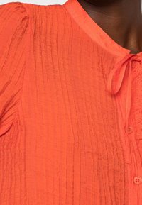 Bright orange shirt featuring a textured, crinkled fabric, a round neckline, a tied detail, and a panel with button accents.