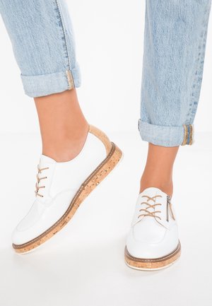 Hvide snøresko med korksåler båret sammen med opkrogede lyseblå jeans på en person, der står mod en hvid baggrund.
