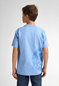 Camiseta de algodón azul claro con mangas cortas, gráfica trasera que presenta un logotipo circular en azul más oscuro, diseño clásico de cuello redondo, corte relajado.